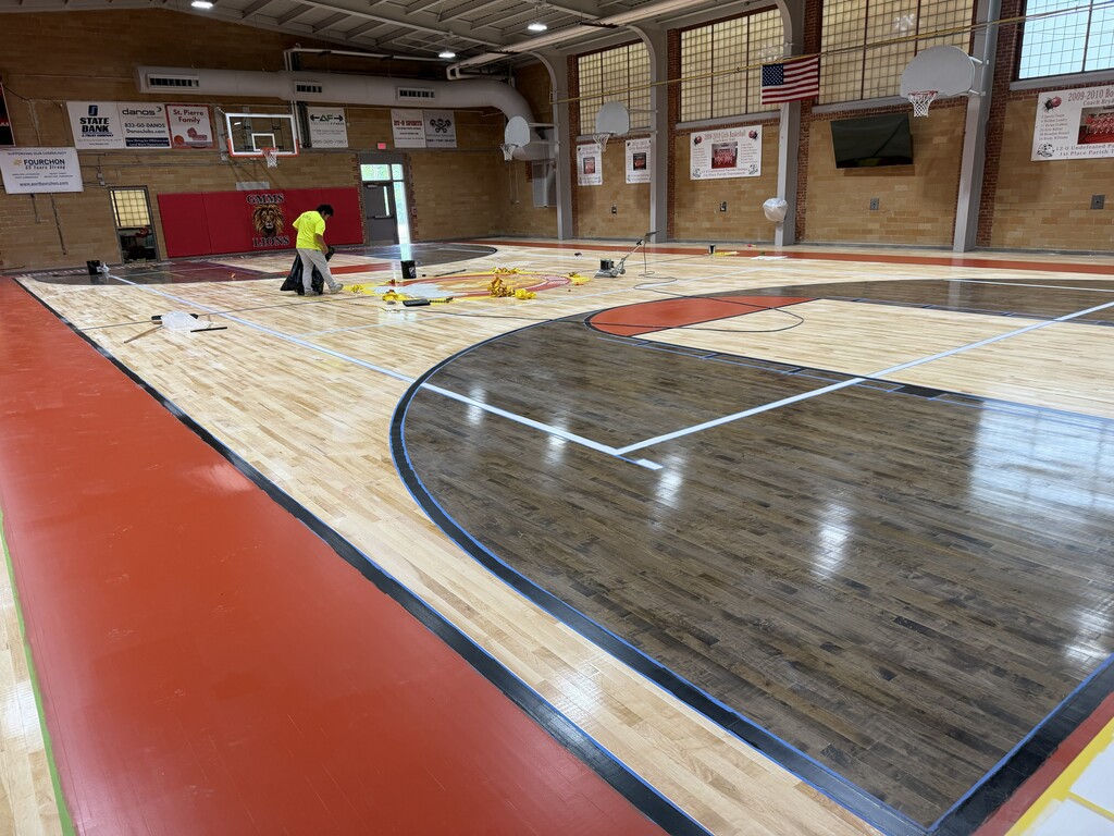 An eye-level, wide-angle photograph taken from the side baseline of an indoor basketball gymnasium showing a floor refinishing project in progress.  The newly polished basketball court features a light-colored natural wood floor contrasted by a dark-stained wood texture inside the three-point arc in the foreground, white court lines, and a wide red painted border. Blue painter's tape lines the edges of the dark-stained section. In the background near the center circle logo, a worker wearing a neon yellow shirt and a dark cap is standing and managing black trash bags and yellow maintenance materials. An industrial floor buffer sits nearby on the court.  The background shows brick walls decorated with various sponsor advertisements and school sports banners, including a 2009-2010 girls' and boys' basketball championship sign. A red wall pad behind the far basket reads "GMMS LIONS" with a lion mascot graphic. An American flag hangs on the far wall next to a mounted flat-screen television.
