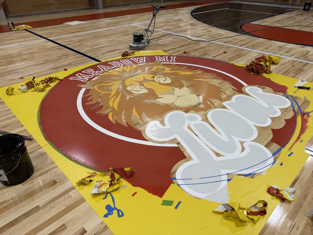 A close-up, high-angle photograph focusing on the center court circle of a basketball gymnasium floor during a painting or refinishing process.  The large circular logo features a detailed illustration of a lion's head with a flowing mane in shades of orange, yellow, and white, set against the natural wood background. The top of the red outer ring reads "MEADOW MI" in white block lettering, and the script word "Lions" is painted in large white letters outlined in blue across the bottom right.  The area surrounding the logo is covered in bright yellow adhesive stenciling sheets, some of which have been peeled back and torn away, leaving crumpled scraps of red, yellow, and blue tape scattered on the polished wood floor. A black paint can sits in the lower-left corner, and an industrial floor buffer with its power cord trailing is parked further back on the court.