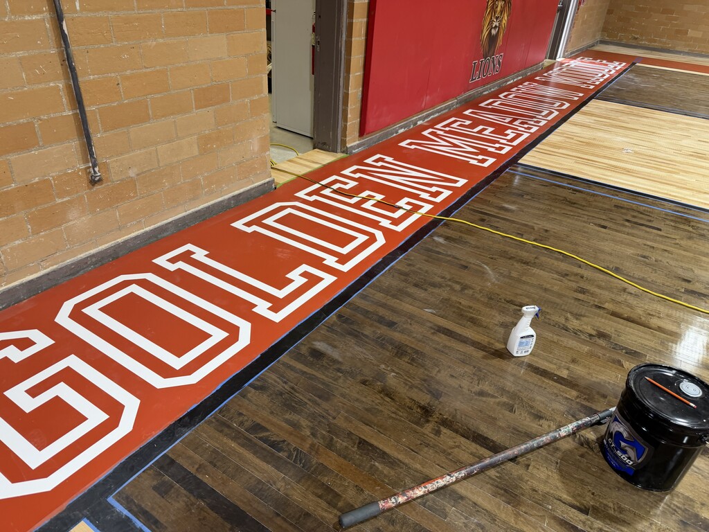 A high-angle, close-up photograph showcasing the red perimeter sideline border of a gymnasium basketball court during a refinishing project.  The wide red border features the words "GOLDEN MEADOW MIDDLE" painted in large, white, sans-serif block letters with a white outline. The border is sharply outlined with blue painter's tape separating it from the adjacent court flooring. To the right of the border, the court transitions into a dark-stained wood floor section where a white spray bottle, a black product bucket, and a long applicator handle lie nearby. A thin yellow extension cord stretches across the floor.  In the background, a light brown brick wall meets a red wall padding that displays a partial lion mascot logo and the word "LIONS".