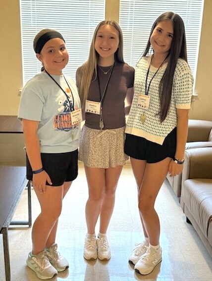 A medium full-length photograph of three smiling teenage girls standing close together side-by-side indoors. They are all looking directly forward at the camera and wearing white name tag lanyards around their necks.  From left to right:  The first girl is wearing a wide black headband, a light blue graphic t-shirt, black athletic shorts, a blue wristband, and grey athletic sneakers. Her name tag reads "Alexandra".  The second girl has long, straight light brown hair and is wearing a dark grey short-sleeve top, a patterned light-grey flared skirt, and white sneakers. Her name tag reads "Mallory".  The third girl has long straight dark hair and is wearing a white short-sleeve knit top with a blue geometric grid pattern, black shorts, a blue wristband, and white sneakers. Her name tag reads "Sydney".  They are standing on a light-colored tiled floor. The background shows an office or lounge setting with window blinds filtering bright daylight, a dark wood table to the left, and a partial view of a grey couch to the right.