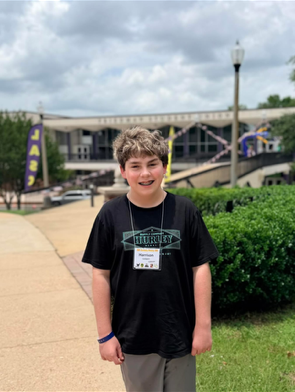 A medium shot of a smiling teenage boy with wavy brown hair standing outdoors on a concrete campus pathway. He is wearing a black graphic t-shirt with a teal diamond logo, grey shorts, a blue wristband, and a lanyard with a white name tag that reads "Harrison".  The background features a slightly blurred view of a large university campus building, the Friedman Student Union, under a cloudy sky. To the right of the pathway are manicured green hedges, and to the left is a tall purple banner. A classic light pole stands prominently behind him to the right.