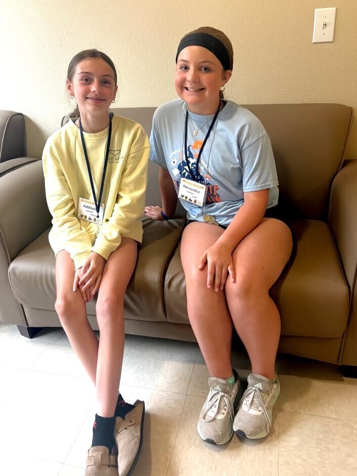 A full-length, eye-level photograph of two smiling young girls sitting side-by-side on a brown leather couch indoors. The girl on the left is wearing a long-sleeve light yellow sweatshirt, black socks, and beige slip-on shoes. She has a lanyard around her neck with a white name tag that reads "Addison". The older girl on the right wears a black headband, a light blue graphic t-shirt, dark shorts, and grey athletic sneakers. She also wears a lanyard with a name tag that reads "Alexandra". They are both looking directly at the camera. The background consists of a plain beige wall with a light switch visible on the upper right, and a light-colored tiled floor is in the foreground.
