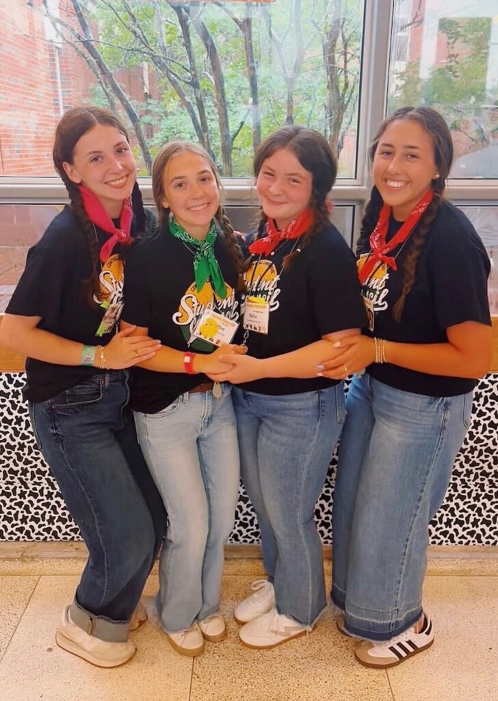 A full-length group photograph of four smiling teenage girls posing closely together indoors in front of a large window.  All four girls are looking directly at the camera, wearing matching black t-shirts with a colorful graphic logo that reads "Student Council," blue jeans, and white sneakers. They each wear a colored bandana tied around their neck and a workshop name tag lanyard.  From left to right:  The first girl has dark hair styled in two braids, wears a pink bandana, dark denim jeans, and leans slightly forward.  The second girl has blonde hair in two braids, wears a green bandana, light-wash jeans, and holds hands with the girl next to her.  The third girl has dark hair in two braids, wears a red bandana, medium-wash jeans, and stands closely linked with her peers.  The fourth girl has long dark hair in a single braid, wears a red bandana, wide-leg jeans, and white sneakers with black stripes.  The background features a window showing green trees outside, and a black-and-white cow print pattern is visible along the base of the wall panel behind them.