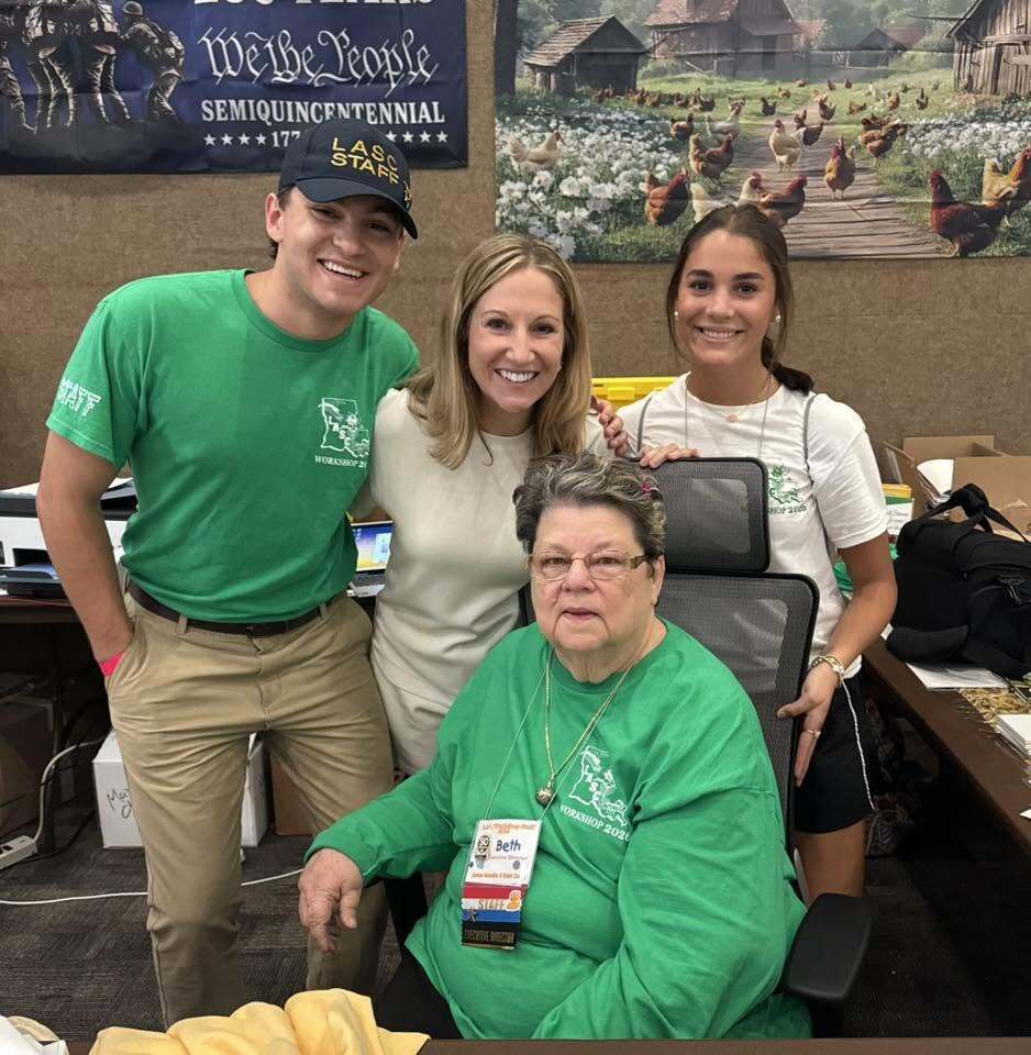 An indoor group photograph of four smiling individuals at a workshop event, positioned around a woman seated in a high-backed office chair. The seated woman has short grey hair, wears glasses, and is dressed in a bright green long-sleeve t-shirt with a "LASC WORKSHOP 2026" graphic logo featuring an outline of Louisiana. She wears multiple credential name tags pinned to her shirt, with the top one reading "Beth." Standing around her are three younger adults: On the left, a young man wears a green t-shirt with the same LASC logo, khakis, and a black baseball cap that reads "LASC STAFF." In the center behind the chair, a blonde woman in a cream-colored top smiles warmly. On the right, a young woman with dark hair tied back in a ponytail wears a white t-shirt with the LASC logo and black athletic shorts. In the background, a dark blue banner reads "We the People SEMIQUINCENTENNIAL," and to the right, a large scenic wall banner depicts an outdoor rural farm landscape filled with chickens. Cluttered office desks with boxes and bags are visible on either side of the group.