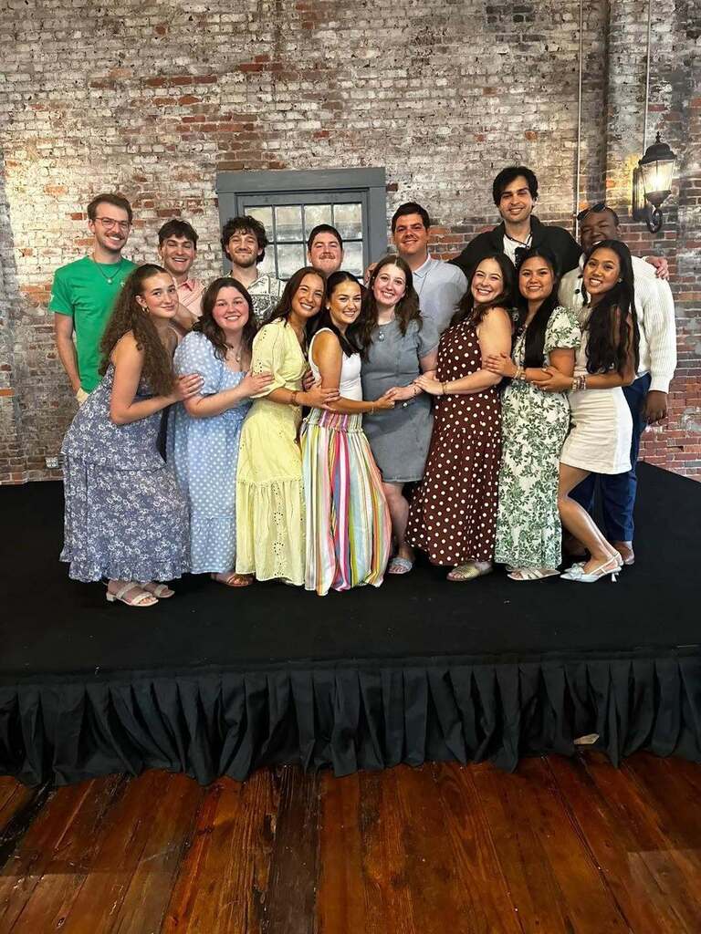 A group photo of fourteen young adult men and women posing closely together on a low, black-skirted stage with a rustic, weathered brick wall background. The group is arranged in two loose rows, smiling warmly at the camera. The women in the front row are dressed in various summer dresses, including blue floral patterns, polka dots, pastel yellow lace, a striped multi-colored maxi skirt with a white tank top, denim, a brown and white polka dot dress, a green floral pattern, and a short white dress. The men in the back row are dressed in casual shirts, including button-downs, t-shirts, and sweaters. The setting features a grey-framed window built into the brick wall behind them, polished hardwood floors in the foreground, and a vintage black lantern-style wall sconce lit up on the far right.