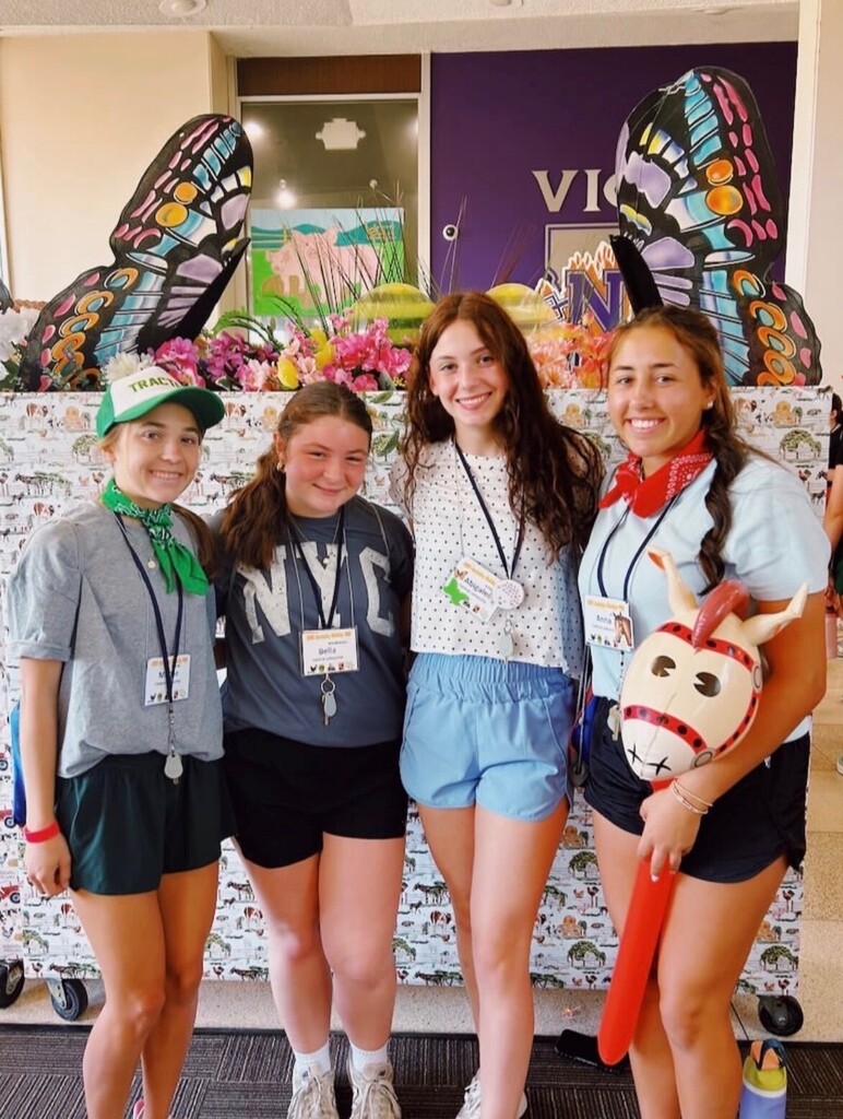 Here is the ADA-compliant alt text for the image image_75ce06.png.  Alt Text: A medium full-length photograph of four smiling teenage girls posing closely together indoors in front of a decorated display table. They are all looking towards the camera and wearing identification lanyards with name tags around their necks.  From left to right:  The first girl is wearing a green and white trucker hat that reads "TRACTOR", a green bandana tied around her neck, a grey t-shirt, dark shorts, and white sneakers.  The second girl has her hair tied back and is wearing a dark grey t-shirt with "NYC" printed in white block letters, black athletic shorts, white crew socks, and beige sneakers.  The third girl has long wavy brown hair and is wearing a white short-sleeve top with black polka dots, light blue athletic shorts, and white sneakers.  The fourth girl is wearing a red bandana around her neck, a light blue t-shirt, black athletic shorts, and holds an inflatable stick horse toy with a cartoon horse face.  Directly behind them is a long table covered in a white cloth patterned with small illustrated farm animal scenes. Atop the table is a colorful spring arrangement featuring pink flowers, green decorations, and two large, upright decorative butterfly wings featuring vibrant patterns in black, blue, yellow, and orange. In the background, a purple wall displays a partial white logo containing the letters "VI" and "N".