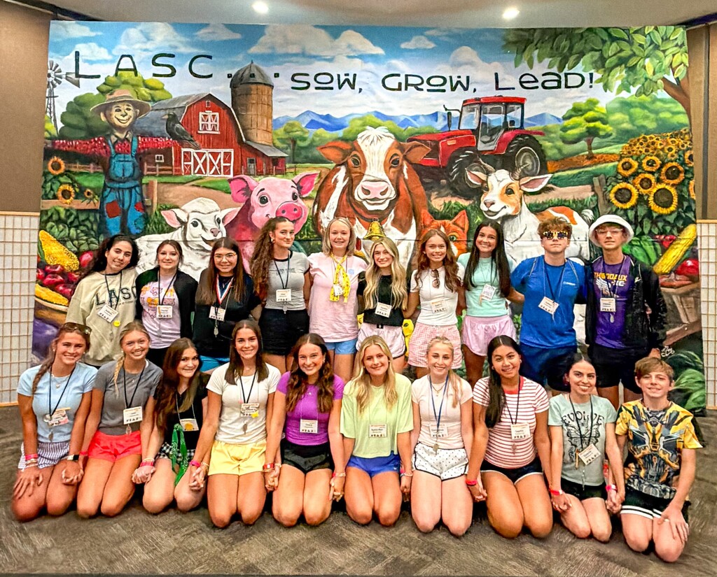 A large group photograph of twenty high school students posing together indoors in front of a colorful painted mural background.  The mural spans the wall behind them, featuring a detailed farm landscape under a blue sky with clouds. The text across the top of the mural reads "LASC... SOW, GROW, LEAD!". The illustration includes a red barn with a silo, a scarecrow, a red tractor, sunflowers, and various smiling cartoon farm animals including a sheep, a pink pig, a brown and white cow, a goat, and a cat.  The students are arranged in two rows:  The front row consists of eleven students kneeling on the grey carpeted floor, holding hands. They are dressed in casual summer clothes including t-shirts, tank tops, and athletic shorts in various bright colors.  The back row consists of nine students standing shoulder-to-shoulder, wearing t-shirts, shorts, and name tag lanyards around their necks. One student on the far right wears a purple shirt that reads "THIBODAUX".  The entire group is smiling and looking directly forward toward the camera.