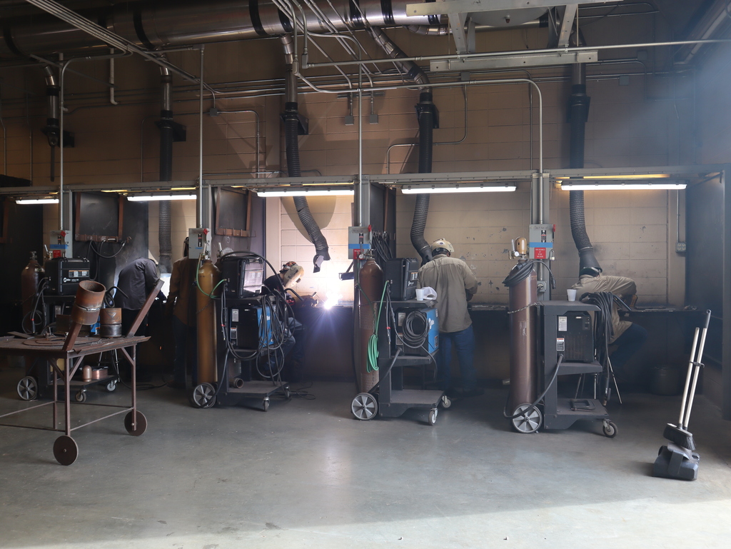 A horizontal, wide-angle photograph capturing a spacious row of five industrial welding stations aligned along a wall in a technical training workshop.  Several students are positioned inside their individual booths with their backs to the camera, working at their respective workbenches. In the middle booth, an intense, brilliant white and blue flare of welding light bursts outward, illuminating the center of the room. The other students are in various stages of preparing or working in darker booths.  In the foreground on the concrete floor, a series of mobile rolling carts are spaced out in front of the stations. Each cart holds industrial welding machinery, thick wrapped cables, and heavy, vertical brown gas cylinders. To the far left, a metal utility table holds scraps of iron pipe, and to the far right, a broom and dustpan rest against the wall. Near the ceiling, an intricate network of metal pipes and thick, black corrugated exhaust duct tubes run down vertically into each booth to vent fumes under fluorescent strip lighting.