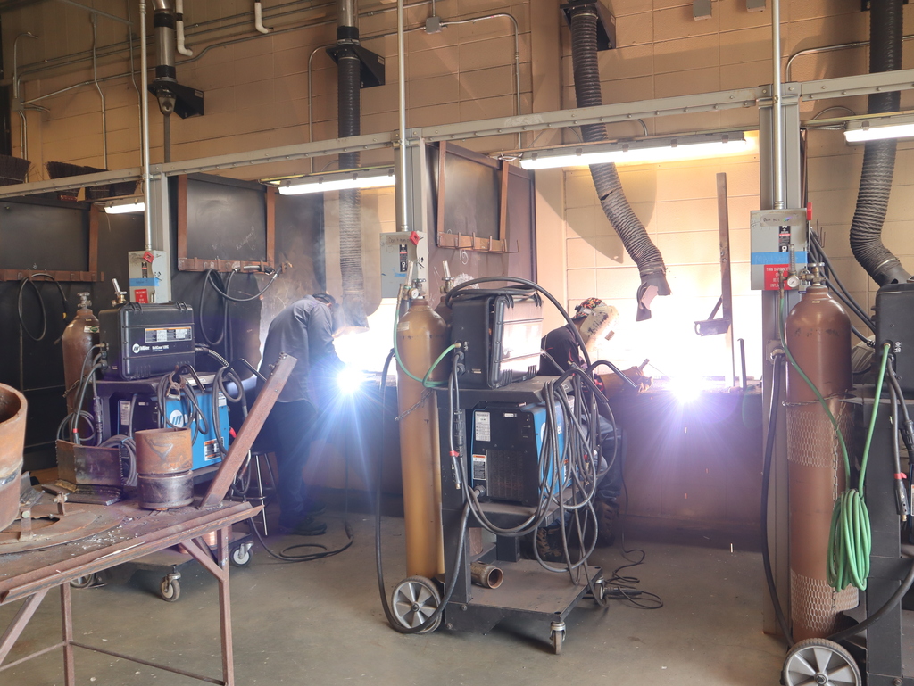 A horizontal, wide-angle photograph capturing a row of industrial welding stations in a high school or technical workshop.  Two students are actively welding in separate, adjacent booths. On the left, a student in a dark long-sleeve shirt stands facing away from the camera, hunched over their workbench. To their right, a second student is seated or leaning over their workstation. Both students are obscured by extremely bright, brilliant bursts of white and blue welding light that create powerful lens flares extending across the booths. Smoke gathers near the ceiling above their stations.  In the foreground, several rolling industrial carts hold welding machines, heavy gas cylinders (including one tan and one brown cylinder with mesh netting), and wrapped black and green hoses. Overhead, fluorescent lights illuminate each individual booth, and thick, black corrugated exhaust duct tubes run vertically up to the ceiling to vent fumes. A metal work table with scraps sits in the lower left corner.