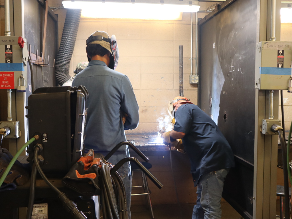 A horizontal, eye-level photograph showing an instructor and a student inside a welding workshop booth.  On the right, an older male instructor wearing a dark blue button-down shirt, blue jeans, protective gloves, and a welding mask leans over a metal workbench. He is actively welding, creating a bright, intense white and blue light that illuminates the center of the frame.  To the left, a student stands with his back to the camera, watching the instructor's demonstration. The student is wearing a light blue denim shirt, jeans, and a welding helmet on his head.  The workshop station features a light-colored concrete block back wall under bright overhead fluorescent lighting. Heavy black vertical divider walls separate the station, and industrial welding equipment and wrapped cables are visible in the dark foreground.