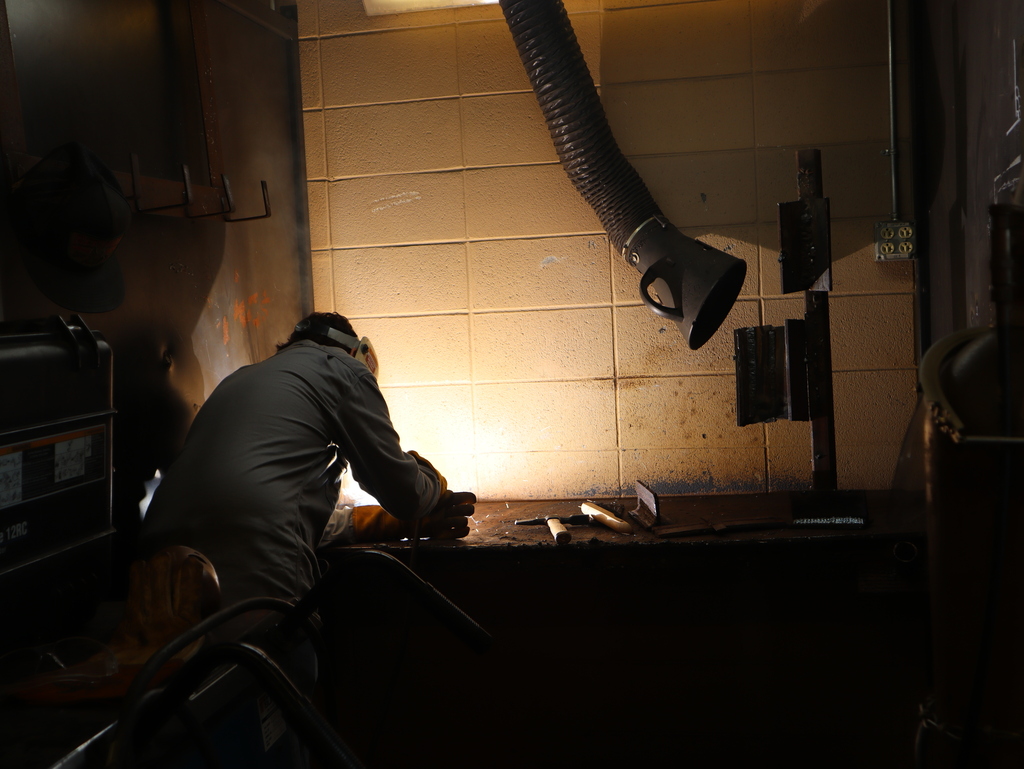 A horizontal photograph of a student welding in a dark shop station.  The student is positioned on the left side of the frame, seen from the back and side, hunched over a metal workbench. They are wearing a long-sleeve grey shirt, heavy protective welding gloves, and a safety helmet. A bright, intense white and golden light glows from the point of welding directly in front of the student, illuminating the concrete block wall behind the workbench.  To the right of the glowing weld, a flexible overhead exhaust vent hose hangs down toward the station. Small metal hand tools rest on the dark surface of the workbench next to a vertical iron welding jig holding metal plates. The rest of the foreground and surrounding workshop booth are cast in dark shadows.