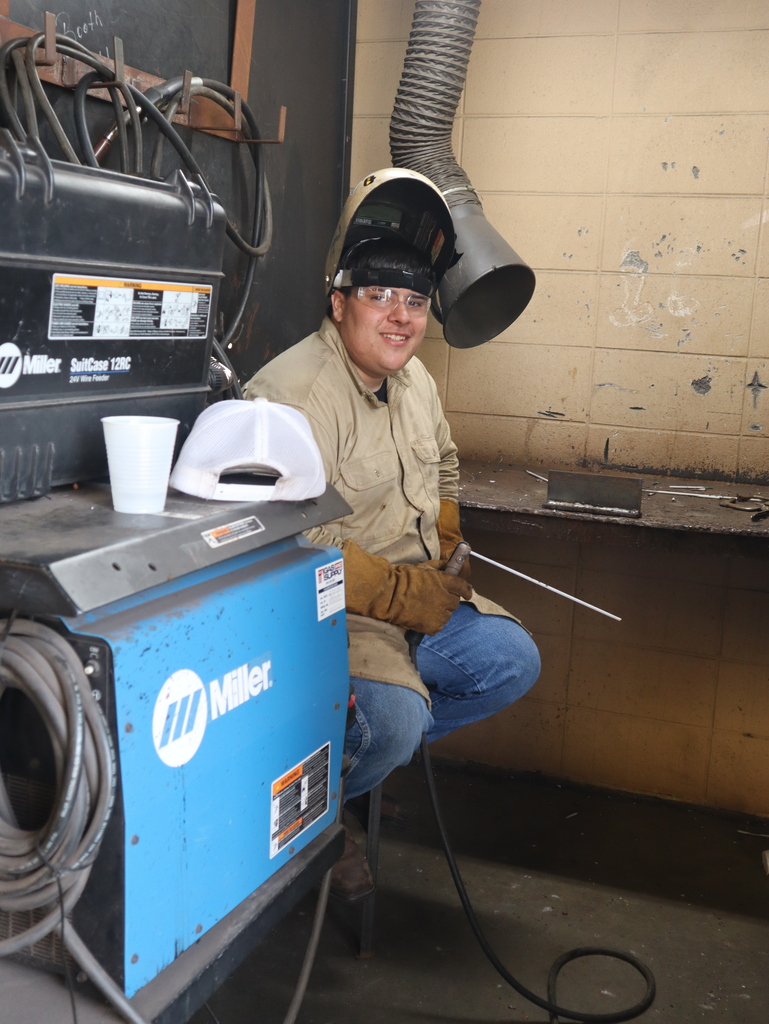 A vertical photograph of a male student smiling toward the camera while seated at a welding workshop station.  The student is sitting in a three-quarter view, looking back over his shoulder. He is wearing clear safety glasses, a tan button-down long-sleeve work shirt, blue jeans, and heavy brown leather welding gloves. His welding helmet is flipped up on his head, and he holds a long, white welding rod attached to a tool handle in his right hand.  In the foreground on the left is a prominent blue and black Miller industrial wire feeder machine with thick cables wrapped around it. A white plastic cup and a white mesh-back trucker hat rest on top of the machine.  The student is positioned in front of a dark metal workbench against a light beige concrete block wall. An overhead corrugated exhaust vent hose hangs down vertically to the right of his head.