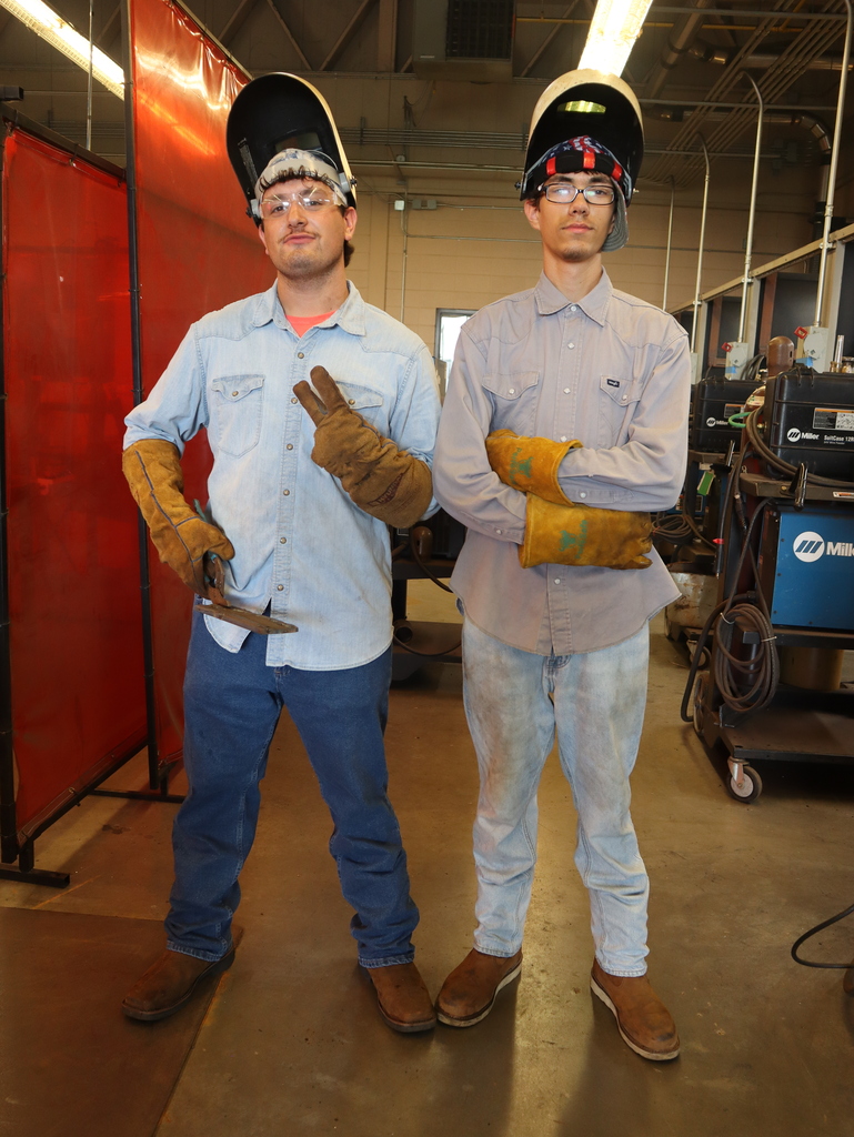 A full-length, eye-level photograph of two male welding students posing side-by-side inside a workshop. They are both looking toward the camera and wearing safety equipment, including clear safety glasses, heavy yellow-brown leather welding gloves, work boots, and welding helmets pushed up on their heads.  The student on the left is wearing a light blue denim button-down shirt, dark blue jeans, and dark brown boots. He stands with his legs slightly apart, holding a flat metal tool in his right hand and flashing a peace sign with his left hand.  The student on the right wears a grey button-down shirt, faded light-wash jeans, and light brown boots. He stands with his arms crossed over his chest.  To the far left is a tall, translucent red plastic welding safety screen. The background features the open concrete floor of the shop, with industrial welding machinery and overhead lighting visible.