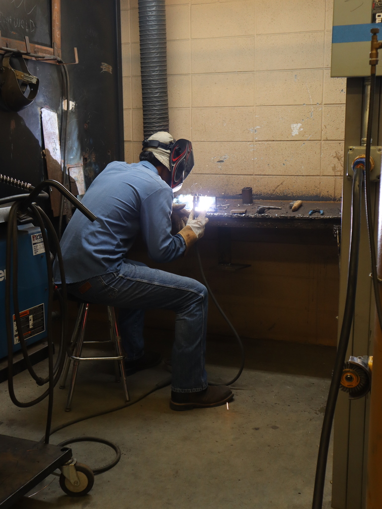 A vertical, eye-level photograph showing a student welding at a workbench inside a workshop station.  The student is positioned on a metal stool, facing away and to the right of the camera. He is wearing a light blue button-down denim shirt, blue jeans, dark work boots, protective welding gloves, and a full welding helmet with graphic detailing. He leans forward over the dark metal workbench as a bright, brilliant white and blue light sparks from the tip of his welding tool.  The station features a concrete block back wall and a large dark metal vertical partition on the left side. A flexible, corrugated ventilation duct hangs down vertically behind the student to collect fumes. Various tools, including pliers and a wire brush, rest on the workbench to the right of the welding area. Elements of industrial shop machinery and cables occupy the dark foreground and borders of the frame.
