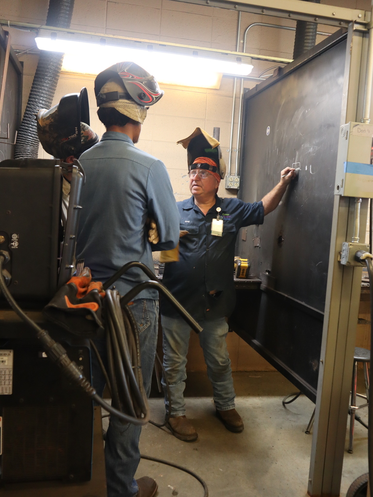 A vertical, eye-level photograph showing an instructor teaching two students inside a welding workshop.  An older male instructor, wearing glasses, a dark blue button-down short-sleeve shirt with a name badge, blue jeans, work boots, and a welding helmet pushed up on his head, stands on the right. He is writing a small diagram on a large black vertical metal divider panel with a piece of chalk.  In the foreground and center, two students stand with their backs mostly to the camera, listening to the instructor. The student in the center wears a light blue button-down denim shirt, blue jeans, brown work boots, and a protective welding mask on his head. To his left, another student is partially visible, also wearing a welding mask.  The workshop environment features concrete flooring, exposed brick and block walls, overhead fluorescent lighting, and welding equipment visible in the lower-left foreground.