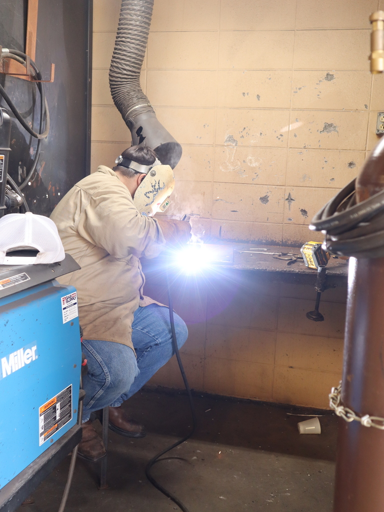 A vertical, medium shot of a student actively welding at a workbench inside a workshop booth.  The student is seated on a stool, facing right and hunched forward over a dark metal workbench. He is wearing a tan button-down long-sleeve work shirt, blue jeans, brown work boots, and a protective off-white welding mask with graphic designs on the front. A brilliant, intense white and blue light with a purple lens flare bursts from the point of welding, illuminating the station and generating a small cloud of smoke.  In the lower-left foreground, a blue Miller industrial machine is visible, with a white trucker hat resting on top of it. A thick black cable runs from the student's welding tool down to the concrete floor.  The background features a worn, light-colored concrete block wall. A large, flexible grey exhaust vent hose hangs down vertically above the student's head to capture smoke. A yellow and black DeWalt tool and loose welding rods rest on the bench to the right of the glowing weld.