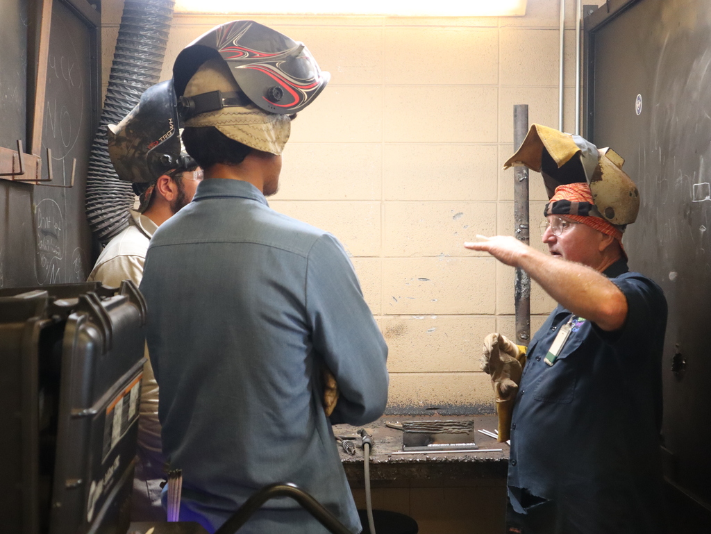 A horizontal, medium close-up photograph of a welding instructor talking to two students inside a workshop station.  On the right, an older male instructor wearing glasses, a dark blue short-sleeve shirt with an ID badge, a colorful orange headwrap, and a welding helmet flipped up on his head is speaking while gesturing horizontally with his raised left arm. He holds a heavy work glove in his right hand.  On the left, two male students stand with their backs mostly toward the camera, listening to the instructor. The student in the foreground wears a light blue long-sleeve button-down shirt and a welding helmet with a decorative red and white flame pattern. To his left, another student is partially visible, wearing safety glasses, a tan shirt, and a dark welding mask.  The background shows a beige concrete block wall under fluorescent lighting, a dark metal workbench with practice welding plates, and a vertical metal pipe. Industrial equipment and partitions frame the left and right sides of the scene.