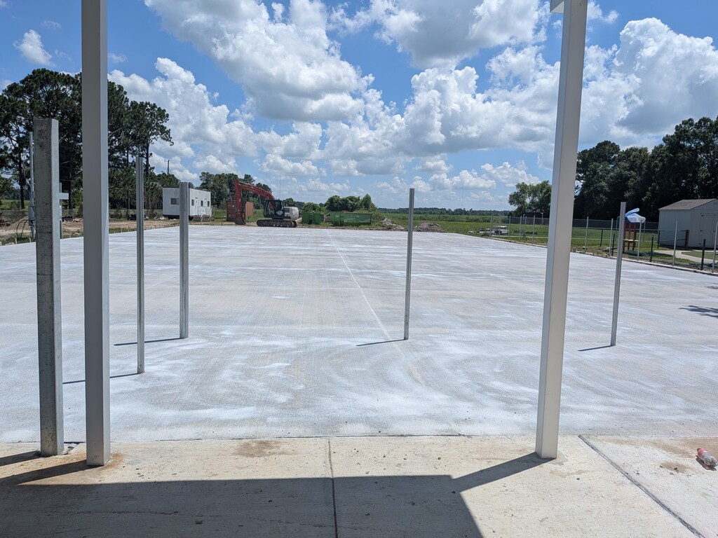 A horizontal, wide-angle photograph showing a large, newly poured flat concrete foundation under a bright, partly cloudy sky.  The perspective is from beneath an open, covered structure, with several thick square white support posts and smaller thin metal poles framing the foreground. The expansive grey concrete pad stretches across the center of the image, showing light surface variations from drying.  In the background, the edge of the construction site meets a grassy green field. An orange excavator, a small white mobile office trailer, a green dumpster box, and a small storage shed are visible along the perimeter of the field. Lush green trees line the far horizon under a vibrant blue sky filled with fluffy white cumulus clouds.