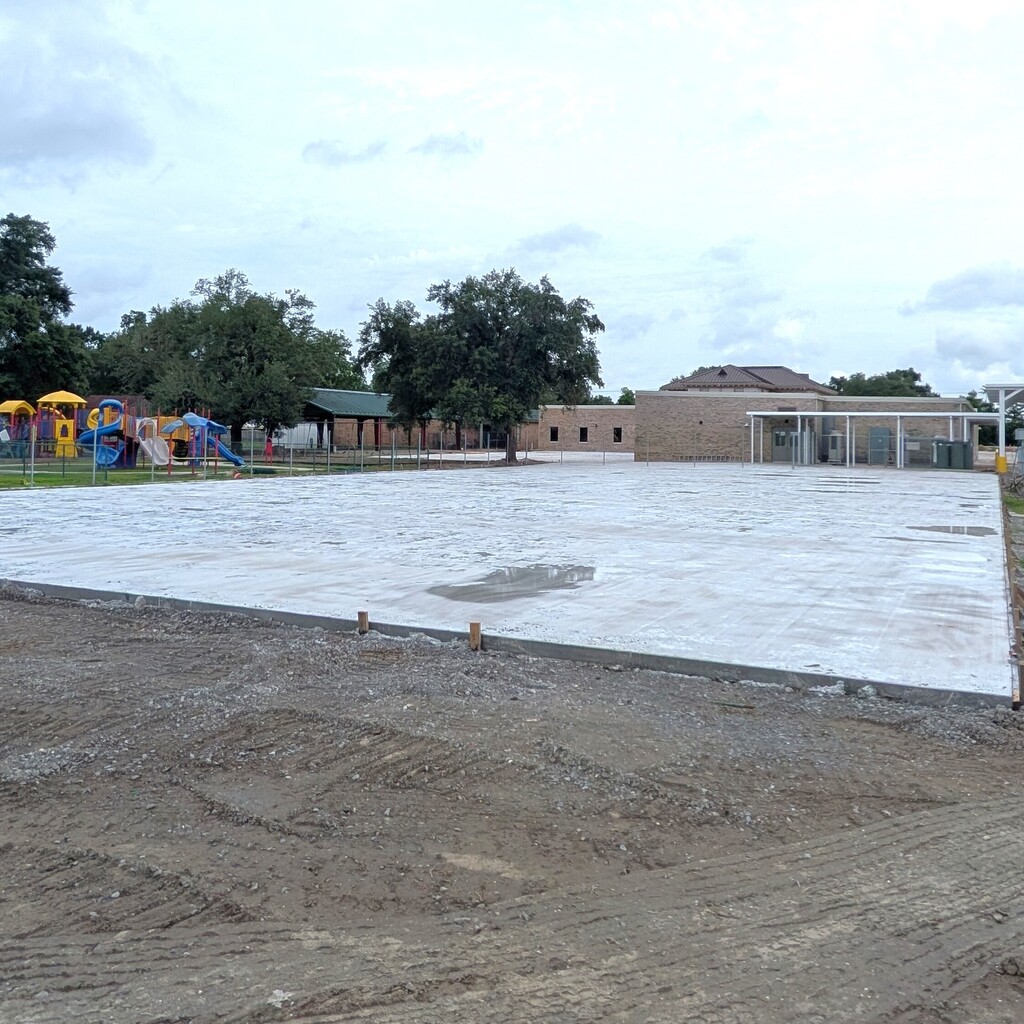 A horizontal, eye-level photograph showing a wide view of a large, newly poured rectangular concrete slab on a school campus.  The concrete foundation is located in the midground, with a light puddle of water reflecting on its surface. The foreground consists of rough, unpaved dark dirt and gravel ground showing faint tire tracks.  In the background on the left, a colorful school playground with yellow roofs and blue slides is enclosed by a fence next to green trees and a covered outdoor pavilion. To the right, a tan brick school building stands with an attached white covered walkway. The entire scene is set under an overcast, bright white and cloudy sky.