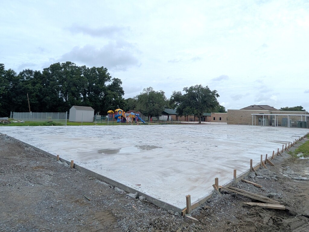 A wide-angle, eye-level photograph of a large, newly poured rectangular concrete slab at an outdoor construction site, likely on a school campus.  The concrete foundation dominates the center of the frame and is contained by wooden forms supported by small wooden stakes driven into the surrounding gravel and dirt ground. A small puddle of water reflects the sky on the slab's surface.  In the background, a school playground with colorful blue, yellow, and red slides and equipment is visible behind a chain-link fence. To the left sits a small grey storage shed. To the right stands a brick school building with a covered