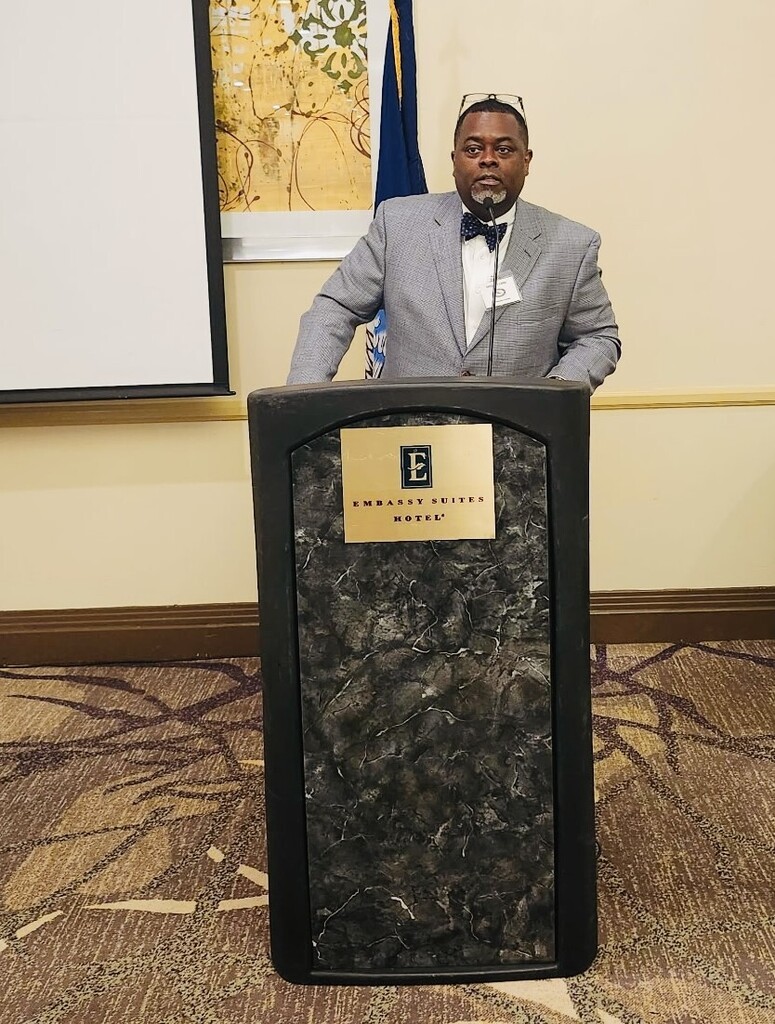 Greg Cook stands speaking behind a tall, dark marbled podium at a conference. He is a Black man with a goatee and glasses resting on top of his head, wearing a grey patterned blazer, a white button-down shirt, and a dark blue polka-dot bowtie. A white name badge is pinned to his jacket pocket, and a microphone is positioned in front of him.  The gold plaque on the front of the podium reads "Embassy Suites Hotel." In the background, a white projector screen stands to the left, a framed abstract painting hangs on the cream-colored wall, and the Louisiana state flag is partially visible behind him. The room has a patterned brown and gold carpet.