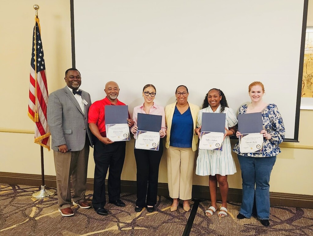 A group photo of six people standing in a line in a conference room in front of a large, blank white projector screen. An American flag stands on a pole to the far left. Four individuals in the group are smiling and holding open blue certificate folders displaying their awards.  From left to right:  Greg Cook, a Black man with a goatee wearing a grey blazer, brown slacks, a white button-down shirt, and a dark bowtie.  A bald man with a grey beard wearing a bright red polo shirt and dark slacks, holding a certificate.  A woman with her hair pulled back wearing glasses, a light pink button-down shirt, and black trousers, holding a certificate.  A woman with glasses wearing a pale yellow blazer over a blue blouse and khaki trousers.  A Black woman with long hair wearing a light-colored striped dress and white sandals, holding a certificate.  A woman with red hair wearing a blue and white floral patterned blouse, blue jeans, and black shoes, holding a certificate.