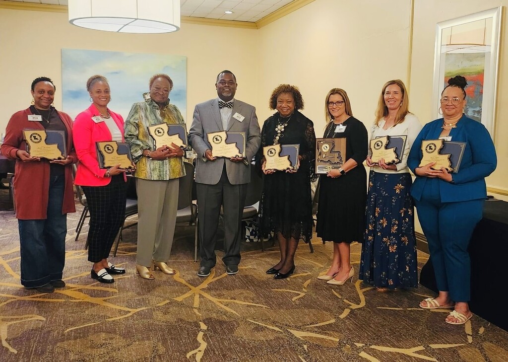 A group photo of eight educators standing in a line in a conference room, each smiling and holding up a professional service award. Most of the awards are plaques shaped like the state outline of Louisiana.  From left to right:  A woman in a long rust-colored cardigan, black top, and dark jeans.  A woman in a bright pink blazer, white top, black checkered pants, and black-and-white loafers.  A woman in a patterned green-and-yellow tunic jacket and khaki slacks.  Greg Cook, wearing a grey blazer, dark slacks, and a striped bowtie, positioned in the center of the group.  A woman in a black lace dress and a statement necklace.  A woman wearing glasses, a black mid-length dress, and nude heels.  A woman in a white long-sleeve blouse and a long, patterned navy blue skirt.  A woman in a teal blue pantsuit and gold sandals.  The room features warm yellow walls, a patterned brown and gold carpet, a large circular ceiling light, and a painting hanging on the wall in the background.