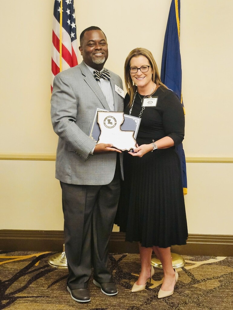 Greg Cook and a female colleague stand smiling side-by-side as they present a service award in front of an American flag and a Louisiana state flag.  Greg, a Black man with a goatee, is wearing a light grey blazer, dark slacks, a button-down shirt, and a striped bowtie. The woman next to him has blonde highlights, wears glasses, a black three-quarter sleeve dress, a silver necklace, and nude heels. Together, they hold up a plaque shaped like the outline of the state of Louisiana, which features a circular LACWAP logo at the top. Both individuals are wearing white conference name badges.