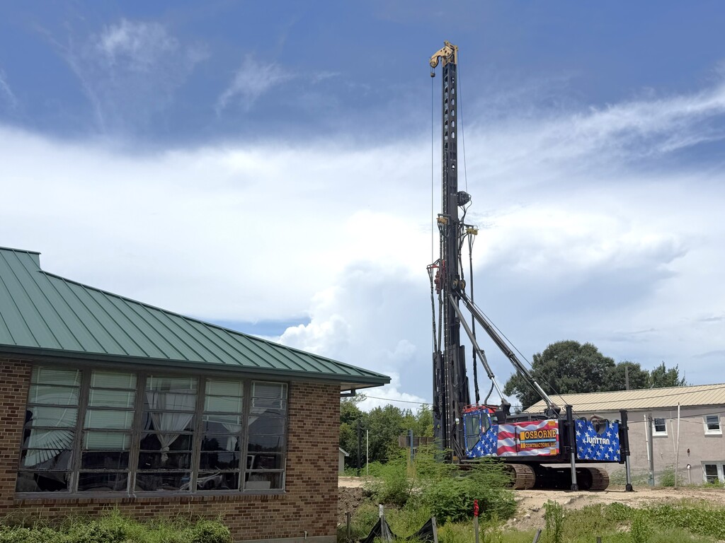 A large Junttan drilling rig wrapped in an American flag pattern with "Osborne Contractors, LLC" branding stands on a construction site next to a single-story brick building with a green metal roof.