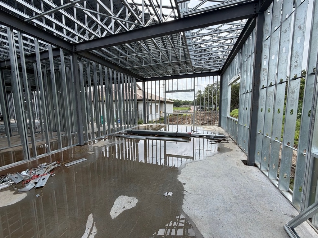 An interior view of a building under construction, showcasing the structural steel framework, open ceiling trusses, and vertical metal wall studs. A large puddle of water covers a significant portion of the smooth concrete floor, creating sharp reflections of the metal framing. In the background, an existing white building is visible through the unsealed wall frames under an overcast sky.
