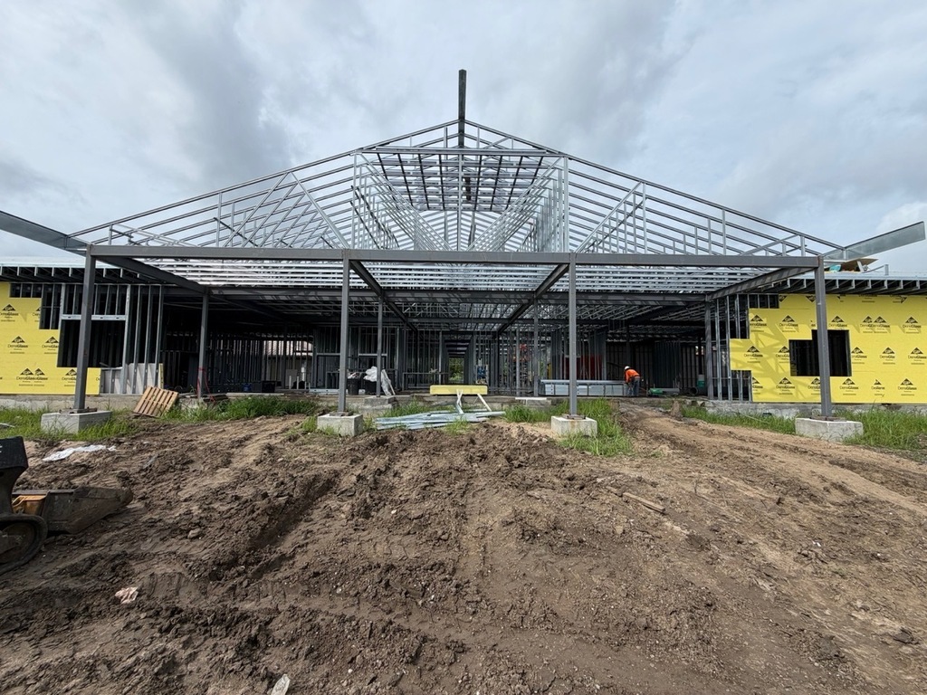 A front-facing view of a large commercial building under construction, featuring a prominent, unroofed peak showcasing its intricate light-gauge steel roof truss system. The structural metal wall framing is visible inside, while the flanking exterior walls are partially covered in yellow Georgia-Pacific DensGlass sheathing. In the foreground, the ground consists of heavily rutted, muddy dirt with a construction worker visible in the distance beneath the frame.