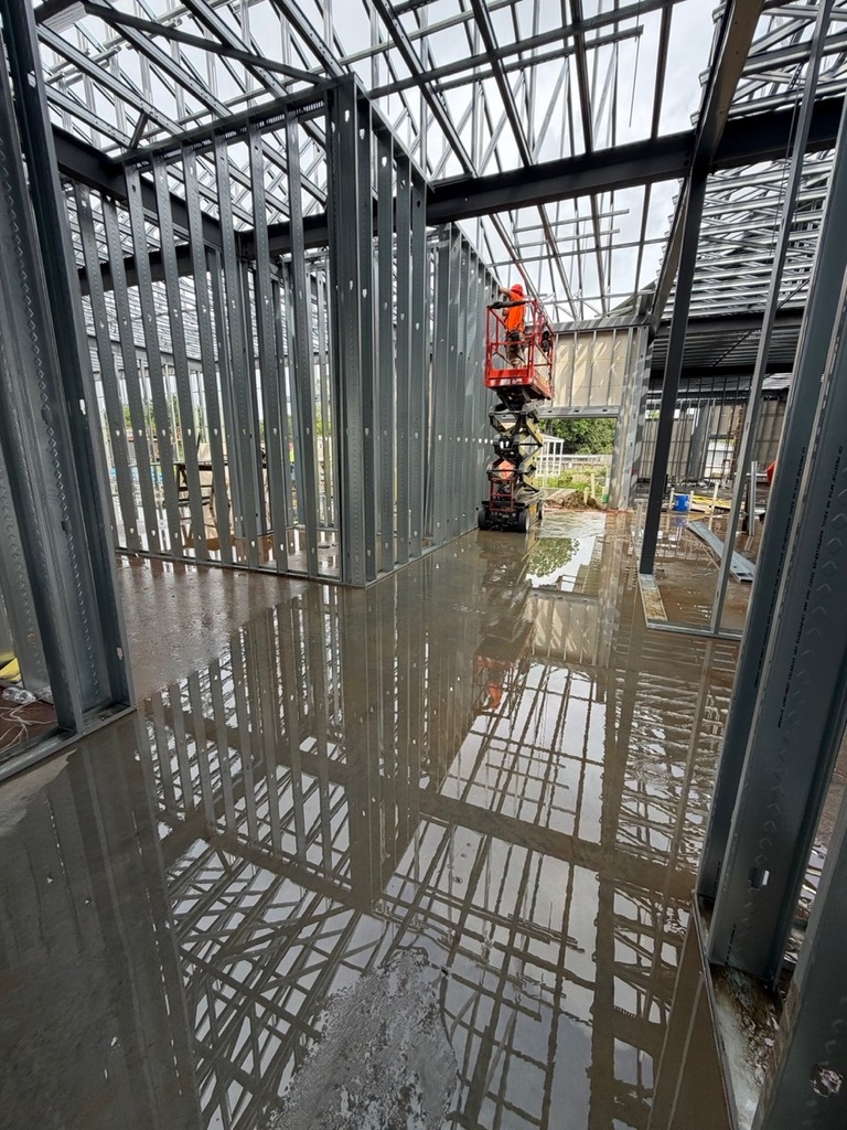 An interior construction photo of a building under progress, featuring exposed metal roof trusses and wall framing. A construction worker in a hard hat is elevated on a red scissor lift, installing materials on an upper level. The concrete floor is wet, creating detailed, distorted reflections of the metal framework.