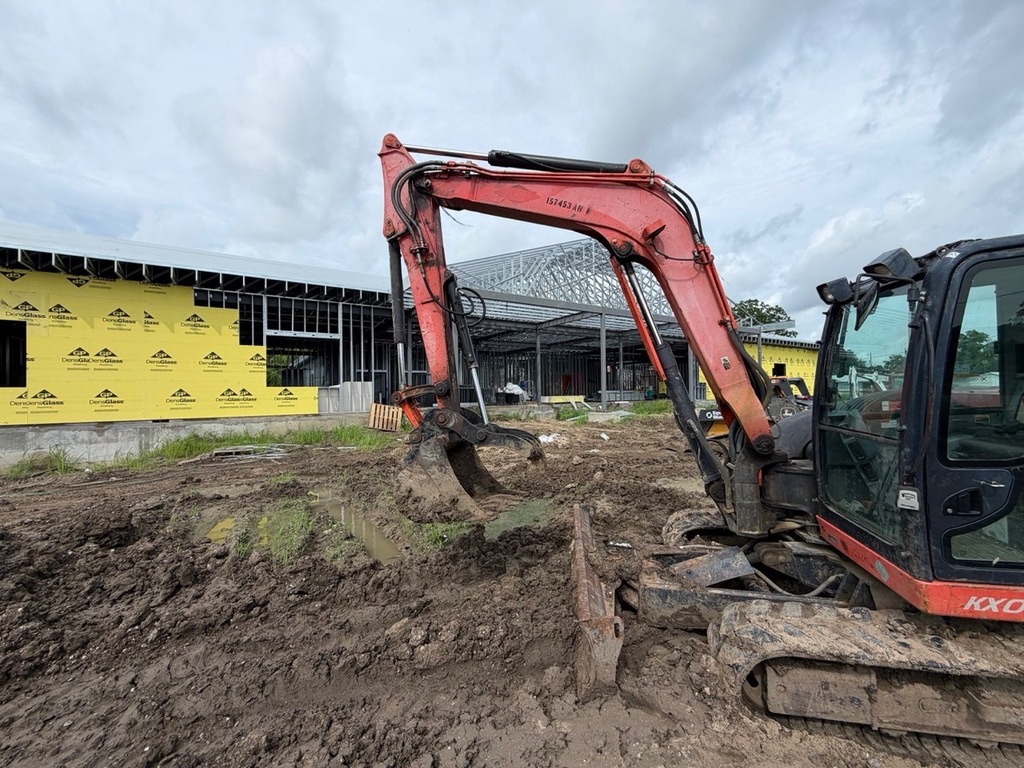 A close-up view of a red and black Kubota KX series excavator parked on a muddy, rutted construction site. In the background, a commercial building is under construction, featuring exposed steel framing and sectioned walls covered in yellow Georgia-Pacific DensGlass sheathing under an overcast sky.