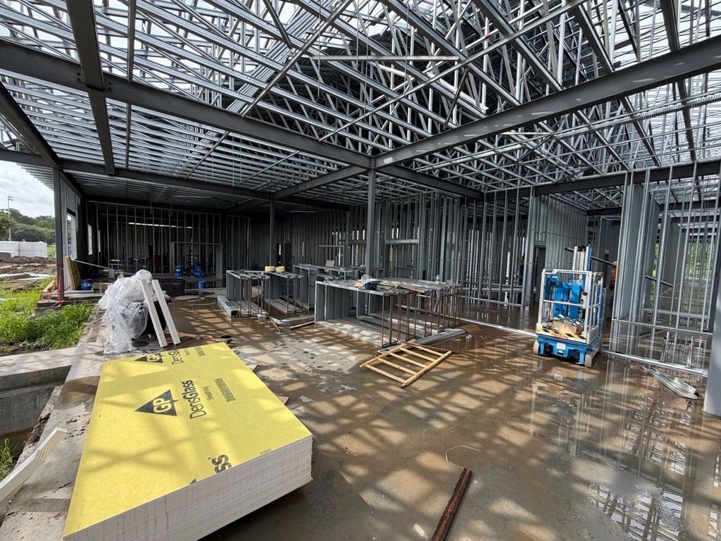 An interior construction site photo shows a large, commercial building under progress, featuring exposed metal roof trusses and wall framing. A pallet of yellow DensGlass sheathing is stacked in the foreground on a concrete slab near a puddle of water. A blue Genie aerial work platform is parked on the right.