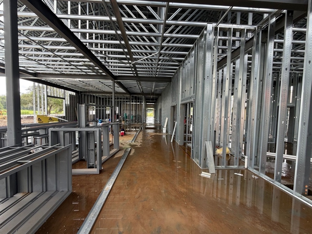 An interior perspective down a long hallway section of a commercial building under construction. The structure features extensive light-gauge steel wall framing, thick structural steel beams, and exposed metal roof trusses. A layer of muddy water covers a large portion of the concrete floor, reflecting the framing, while assembled grey door or window frames are stacked on the left side of the corridor.