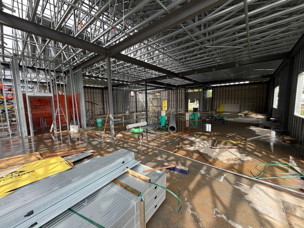 An interior view of a commercial building under construction, showcasing the exposed structural steel frame, metal wall studs, and open roof truss system. Large puddles of water reflect the framing on the concrete floor, which is scattered with construction debris, pipes, hoses, and a push broom. In the foreground, bundles of unused metal framing studs and a piece of yellow DensGlass sheathing rest on the ground.