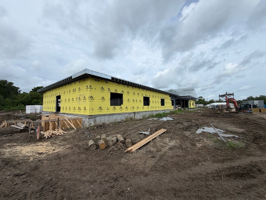 A single-story commercial building under construction with a concrete foundation and walls covered in yellow Georgia-Pacific DensGlass sheathing. The surrounding construction site consists of muddy, uneven dirt with scattered logs, lumber, and puddles, while a red excavator sits parked in the background under a cloudy sky.