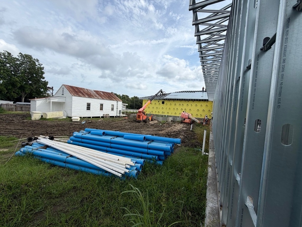 An outdoor construction site with stacks of blue and white PVC pipes on green grass in the foreground. On the right, a wall of vertical metal framing studs extends into the background. In the middle ground, an orange telehandler crane works next to a new building with yellow sheathing, while an old white building with a rusted metal roof stands to the left under a cloudy sky.