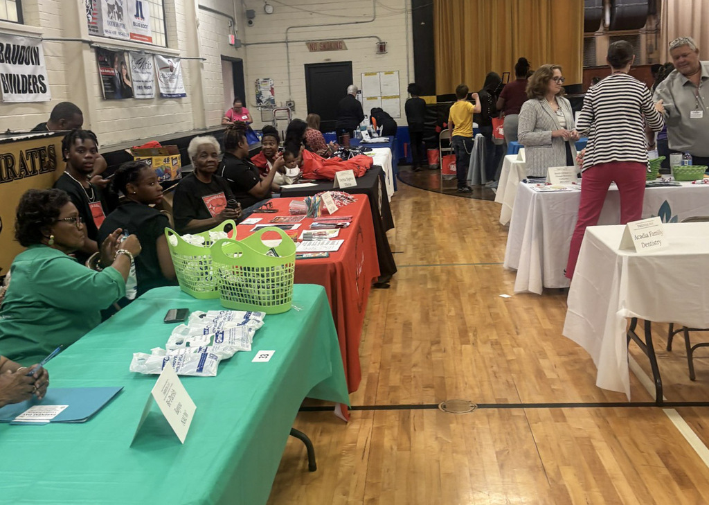 Indoor view of a school gymnasium hosting a community resource fair. Rows of tables with colorful tablecloths are lined up across the wooden gym floor, with community representatives and vendors seated behind display tables featuring flyers, bright green baskets, and promotional materials while attendees walk through.