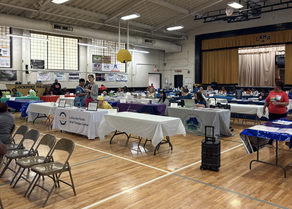Wide-angle view of a community resource fair in the Lockport Middle School gymnasium. Rows of tables covered in tablecloths feature displays for local organizations, including the Lafourche Parish Brief Therapy Center and Lafourche Parish Public Library, with representatives standing and seated throughout the room.