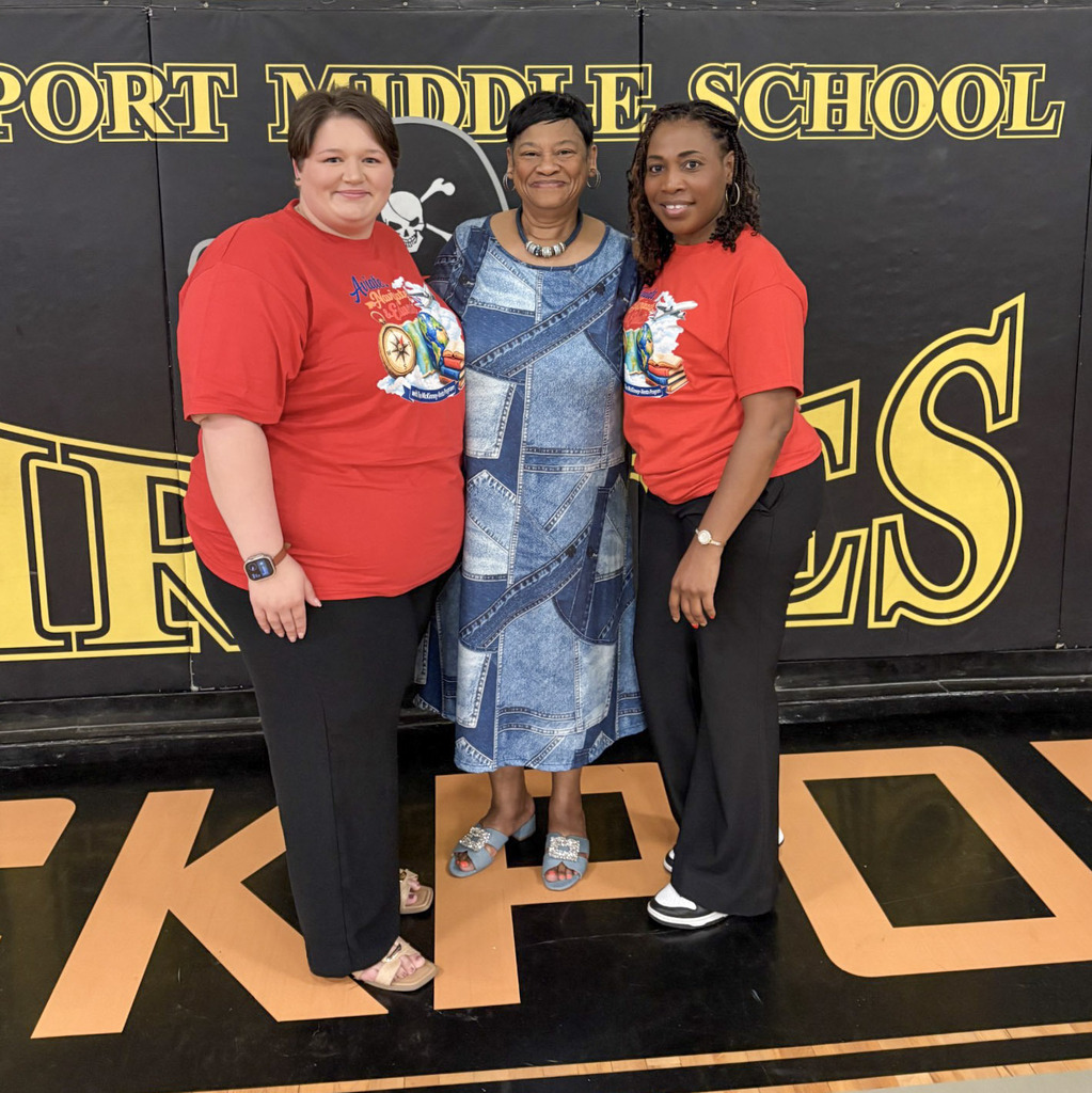 Three women pose together indoors in front of a black and gold Lockport Middle School gym wall. The woman in the center wears a denim-patterned midi dress and blue sandals, flanked by two women in matching red T-shirts and black pants.