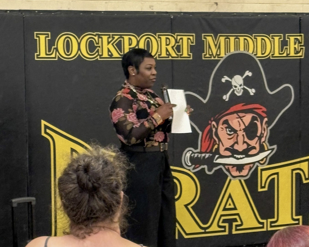 A speaker with short black hair wearing a long-sleeved floral print blouse and dark pants speaks into a microphone while holding papers in front of an audience. She stands in front of a black and gold gym wall featuring the Lockport Middle School pirate mascot logo.