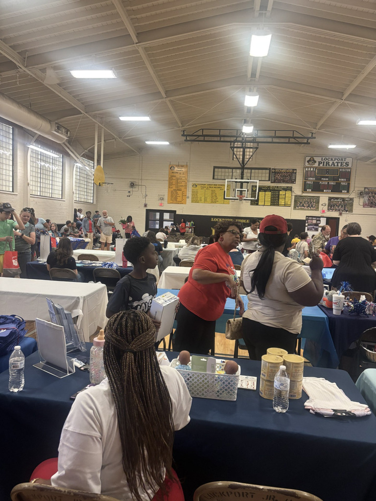 Wide view of a busy community resource fair inside the Lockport Middle School gymnasium. Attendees and family members walk between rows of tables set up with white and blue tablecloths featuring promotional displays, giveaways, infant care items, and educational materials.
