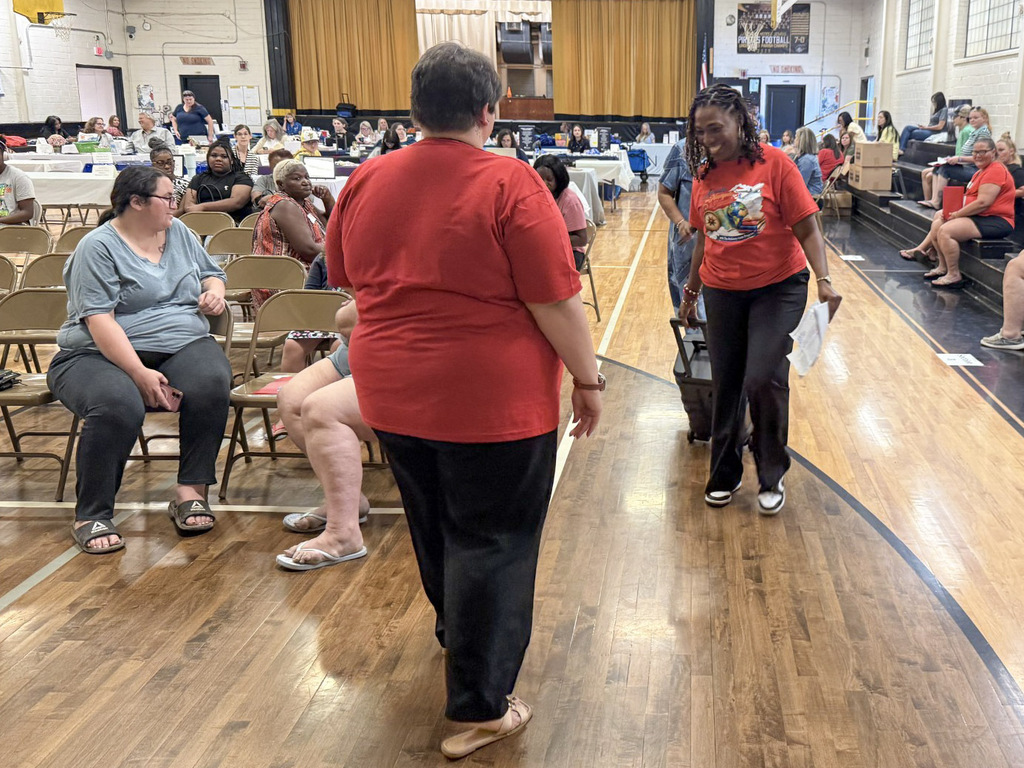 A woman in a red shirt pulls a rolling speaker down the wooden floor of a school gymnasium during an event. A woman in a red shirt walks toward her while seated attendees in metal folding chairs and staff at vendor tables look on.