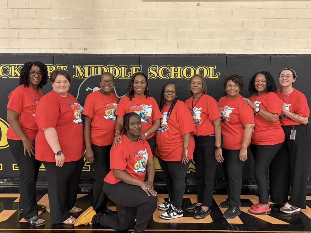 Group photo taken inside a gymnasium in front of a black and gold wall banner that reads "Lockport Middle School." Participants: Nine women, all wearing matching red short-sleeved T-shirts featuring a graphic that reads "Aviate, Navigate, & Educate" with a compass, books, and an airplane graphic. Pose: Eight staff members are standing side-by-side smiling in black pants, with one woman kneeling in the front center in dark pants and bright yellow shoes.