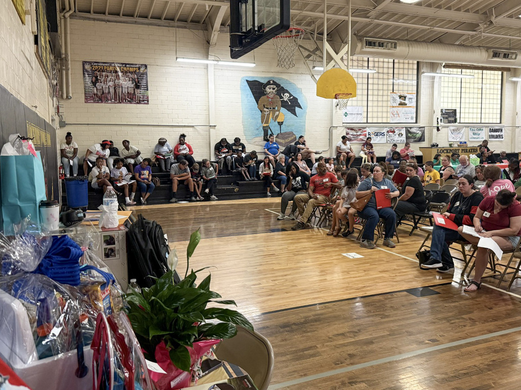 Audience members and families seated in folding chairs and bleachers inside the Lockport Middle School gymnasium during an event. Gift baskets wrapped in cellophane and a potted plant rest on a table in the foreground.