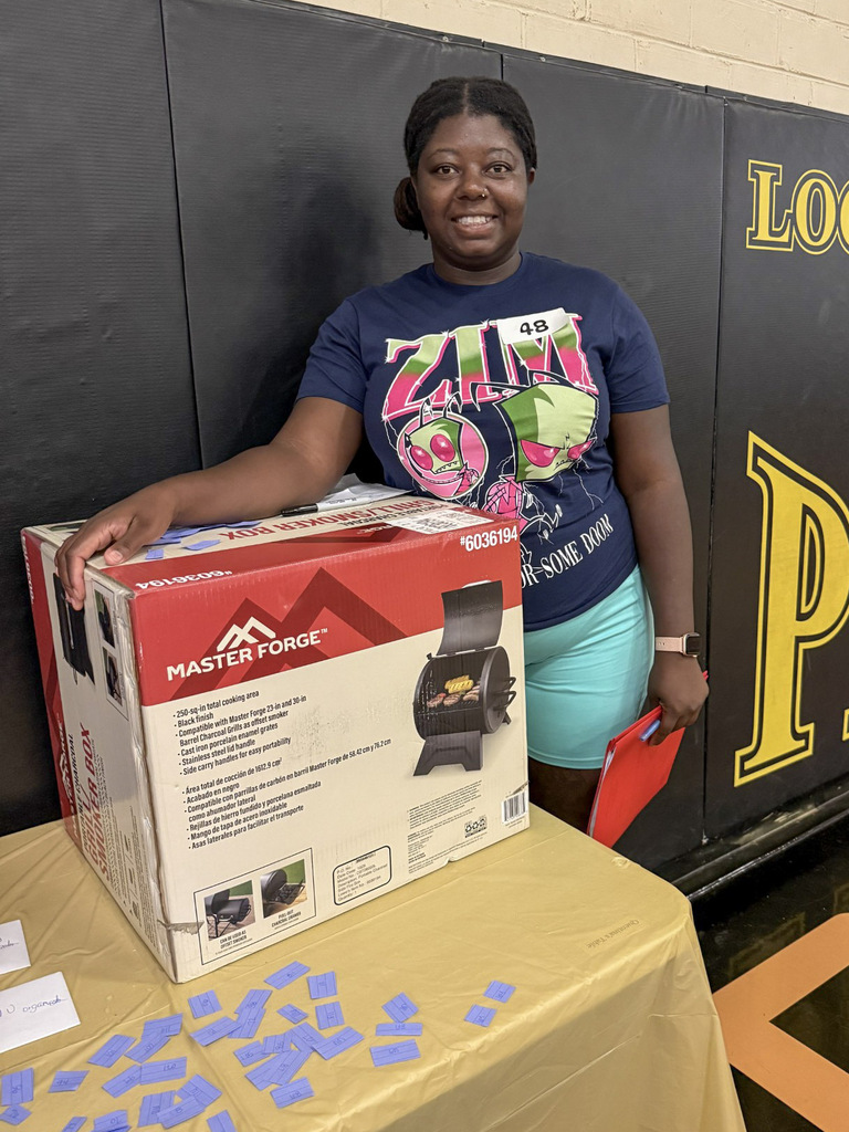 A woman in a navy graphic T-shirt and light blue shorts stands smiling next to a boxed Master Forge portable charcoal grill smoker box resting on a table. Blue raffle tickets are scattered on the table in front of the box.