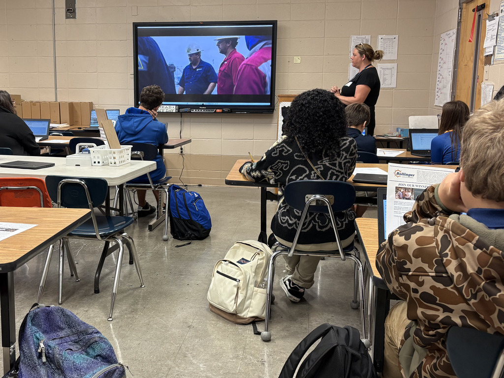 Middle school students sit at individual desks facing a large screen showing a video about industrial shipyard careers, while a guest speaker stands nearby overseeing the classroom presentation.