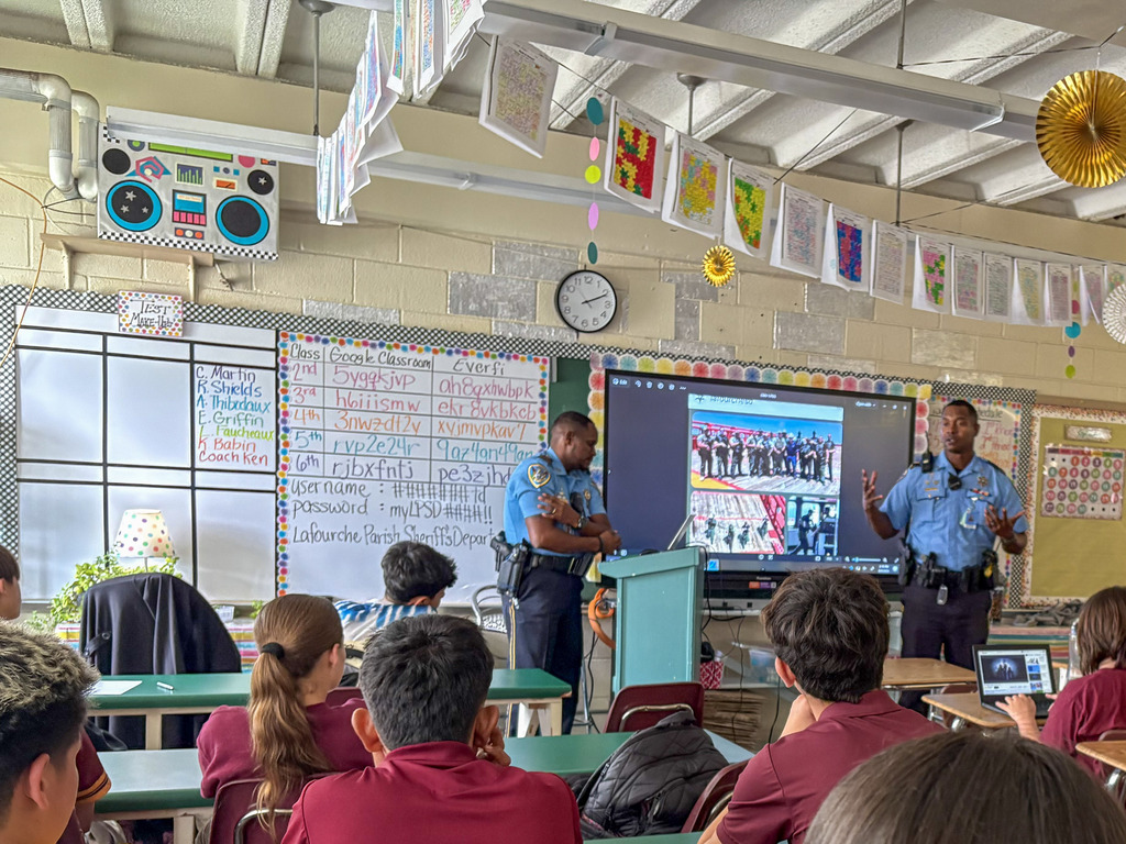 Two Lafourche Parish Sheriff’s deputies in light blue uniforms stand at the front of a middle school classroom, giving a presentation to a group of seated students. A large digital screen behind them displays photos of law enforcement personnel.