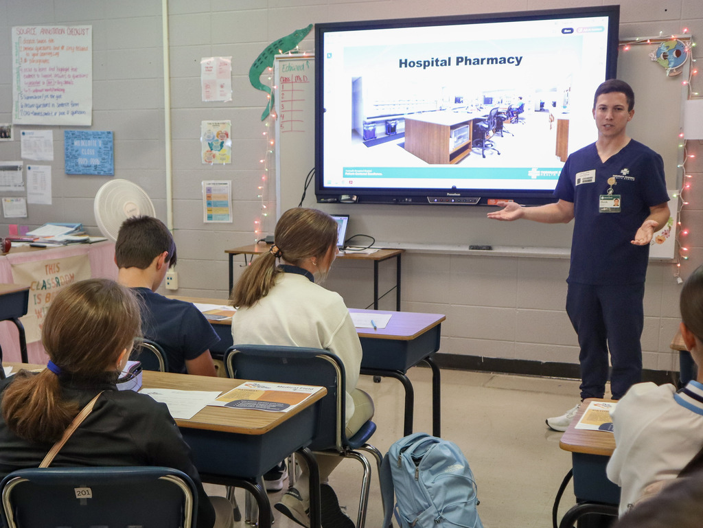 A healthcare professional wearing navy blue scrubs presents to a classroom of students, using a large digital display showing a photo slide titled "Hospital Pharmacy."